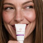 Smiling woman holding freezeframe REVITALEYES 15ml eye gel cream in white packaging, formulated to target deep under-eye wrinkles, chronic puffiness and dark circles.